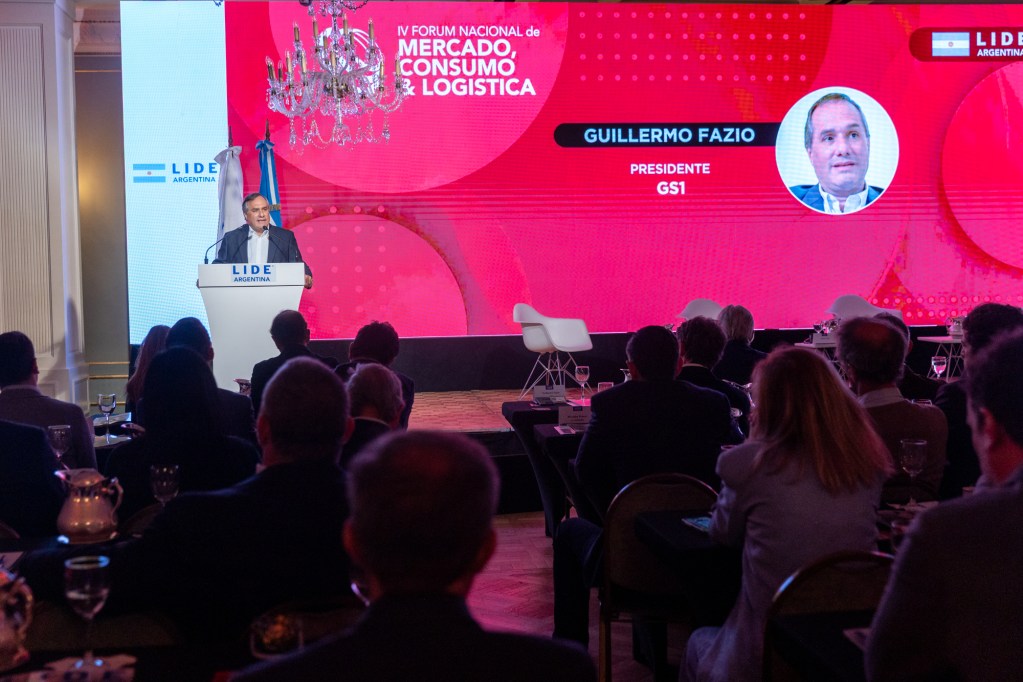Un orador en el IV Foro Nacional de Mercado, Consumo y Logística, con una pantalla de fondo que muestra la información del evento y una foto de Guillermo Fazio, presidente de GS1.