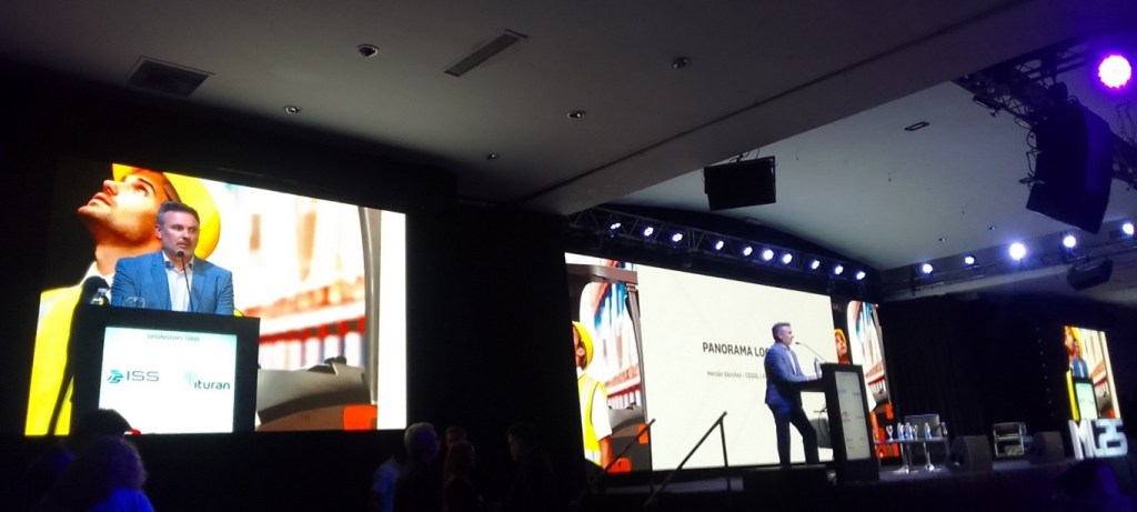 Un orador en un escenario durante el evento Management Logístico 2025, con pantallas en el fondo mostrando gráficos y logotipos de patrocinadores.