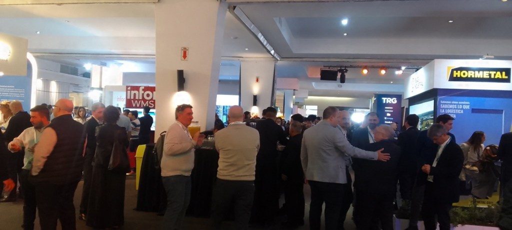 A busy networking event with numerous attendees interacting and engaging at various booths, set in a well-lit indoor venue.