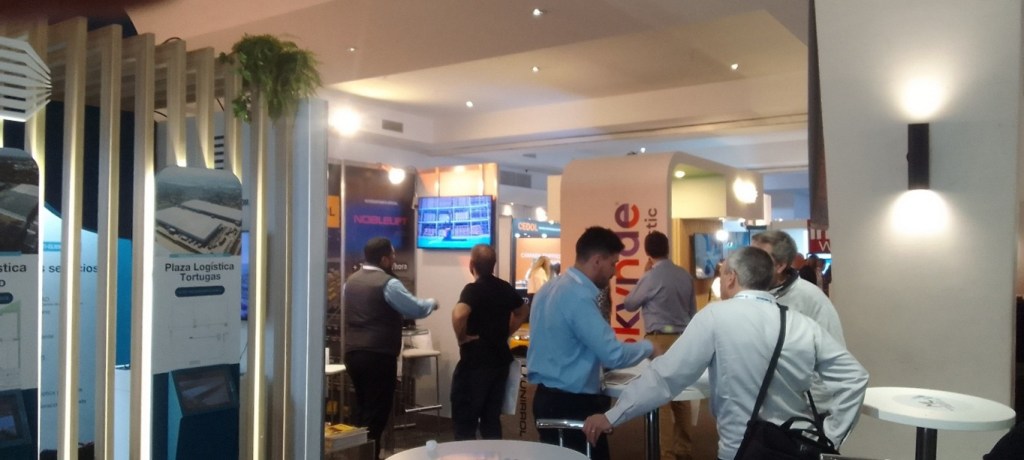 An exhibition space with multiple people interacting, featuring displays and signage related to logistics, showcasing a branded area for 'Plaza Logistica Tortugas'.