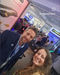 A man and a woman pose for a selfie at a crowded event, with promotional displays and an audience in the background.