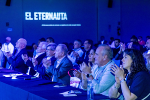 A group of people clapping and sitting at a table during an event, with the title 'EL ETERNATA' projected on a screen in the background.